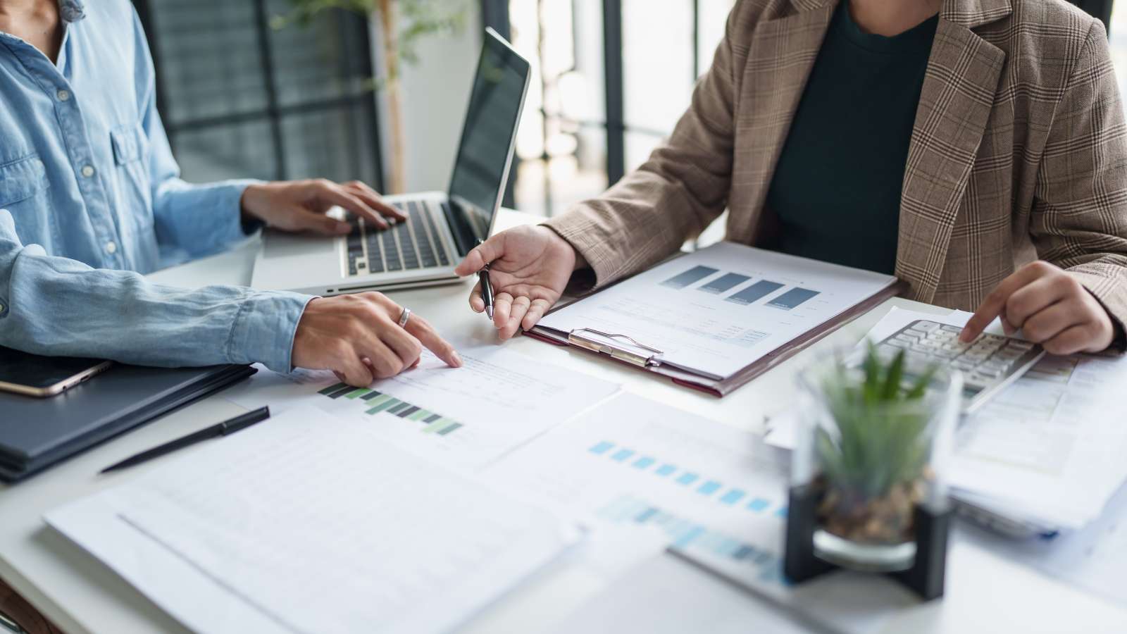Dos personas revisando gráficos y reportes financieros en una reunión de trabajo, con laptop y calculadora