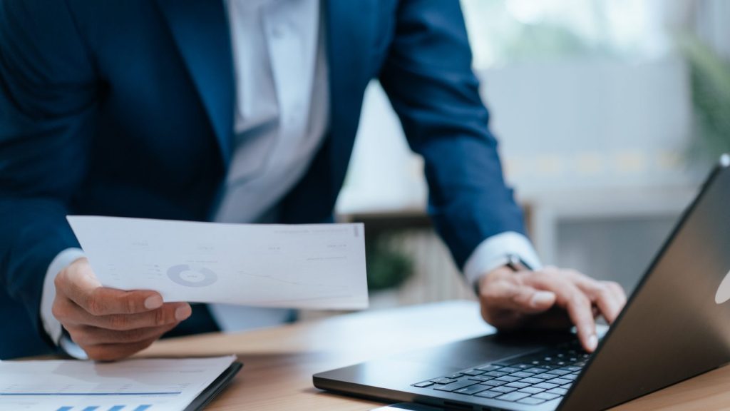 Ejecutivo comparando un informe financiero impreso con su laptop al evaluar una rectificación de renta como empresa