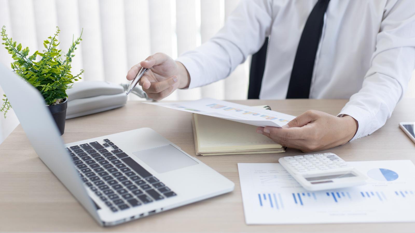Empresario de camisa y corbata revisando gráficos financieros junto a su laptop y calculadora en la oficina