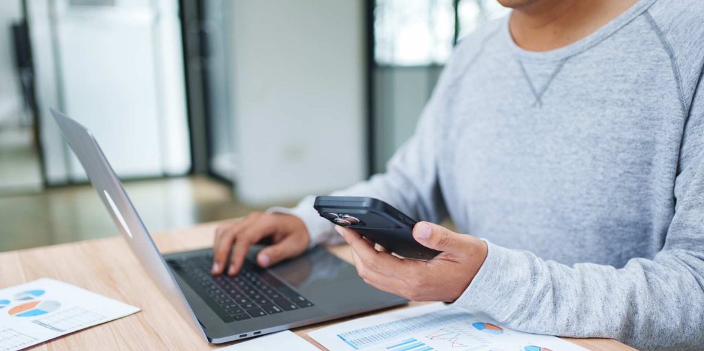 Persona trabajando en un computador portátil con un celular en la mano y informes con gráficos sobre el escritorio para revisar los activos fijos de una empresa